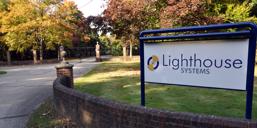 A curved brick wall leading to a wide metal gate surrounded by trees; in the foreground, a sign reading: Lighthouse Systems