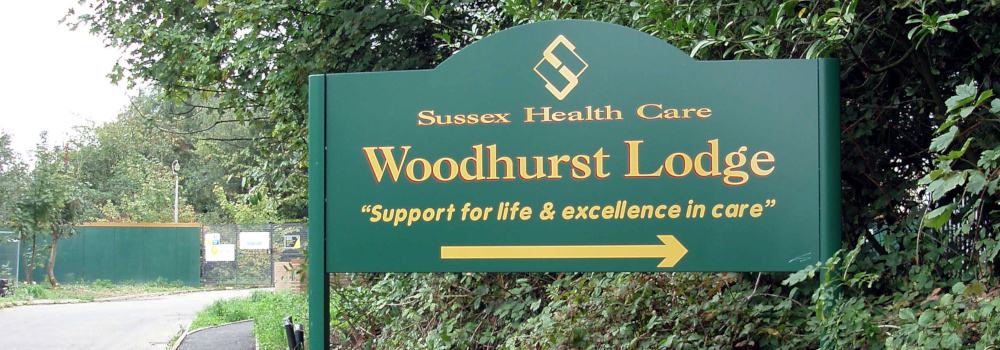 A dark green sign with gold lettering: Sussex Health Care, Woodhurst Lodge, support for life and excellence in care